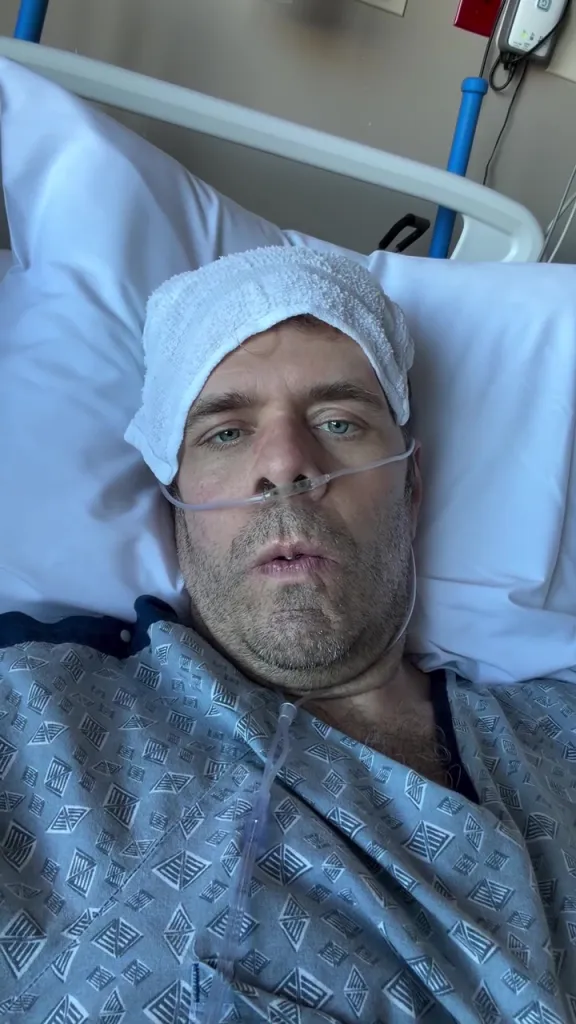 Perez Hilton in a hospital bed with a towel on his head and a nasal cannula.