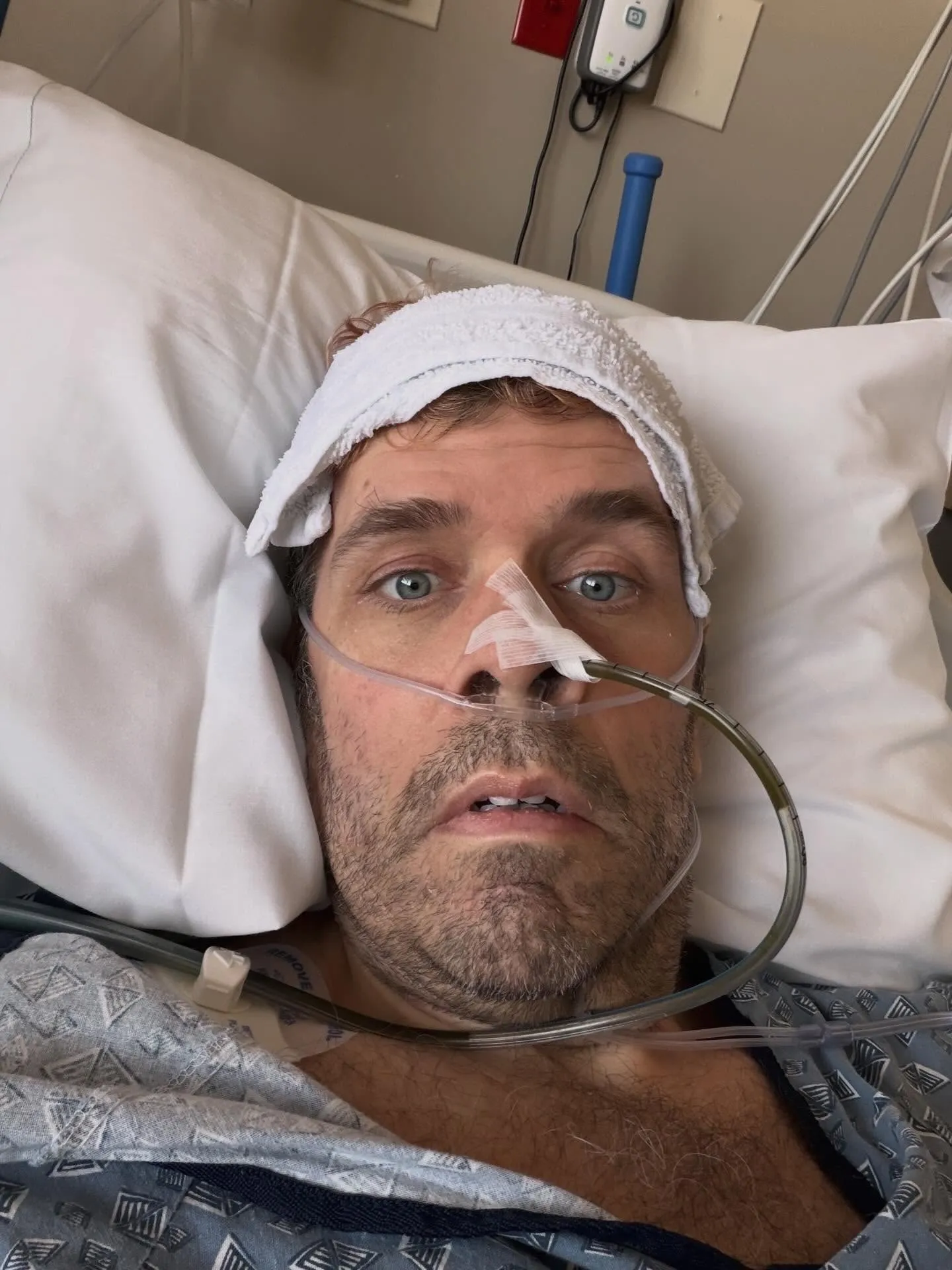 Perez Hilton lying in a hospital bed with a white towel on his forehead and a nasal tube.