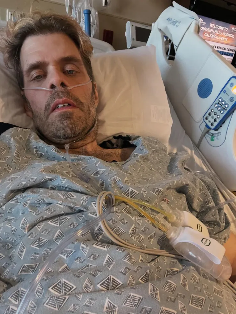 Perez Hilton lying in a hospital bed with a nasal cannula and medical drains attached.