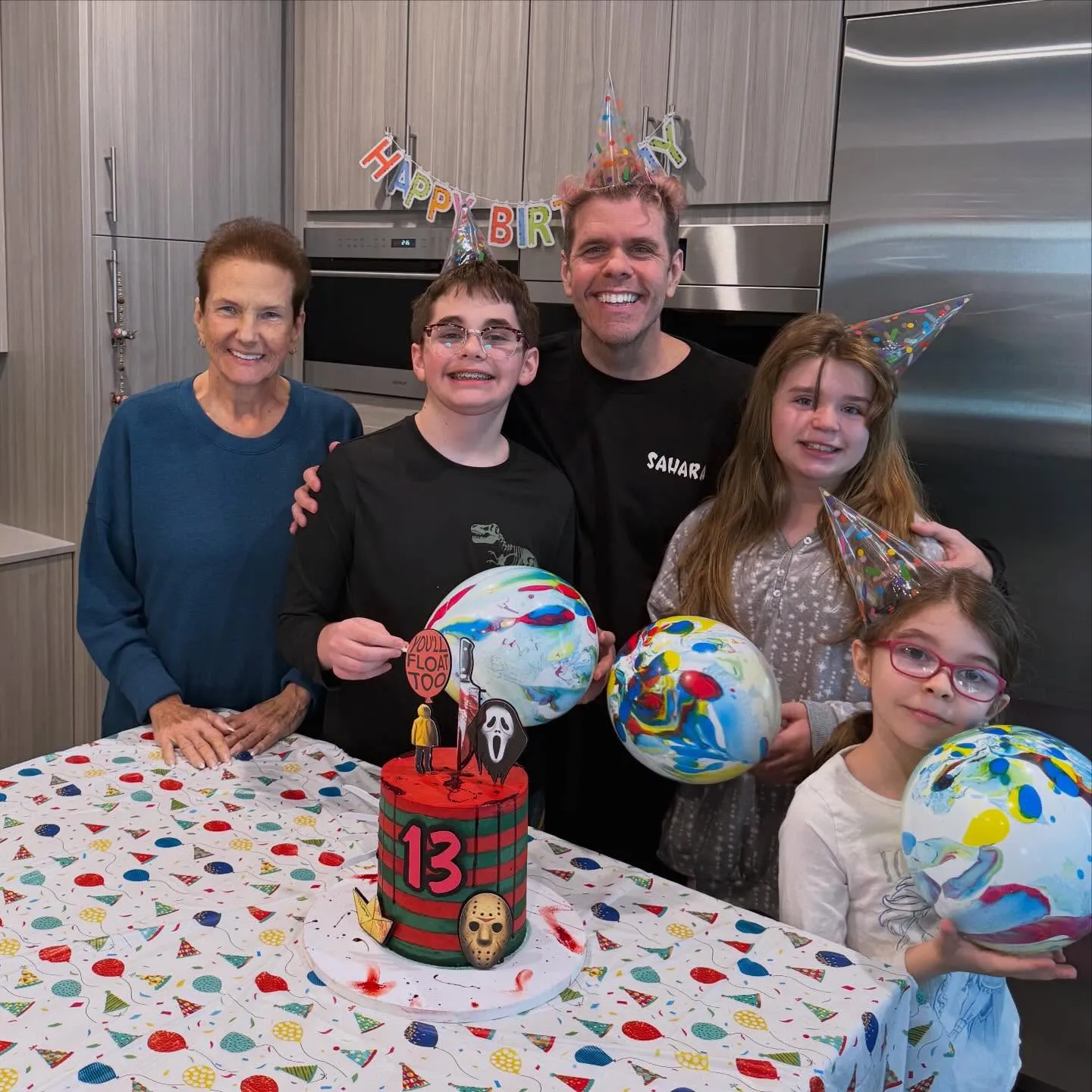 Perez Hilton, his mother, and three children stand behind a birthday cake and balloons.