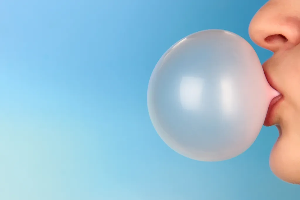 A person blows a large pink bubble with chewing gum against a blue background.