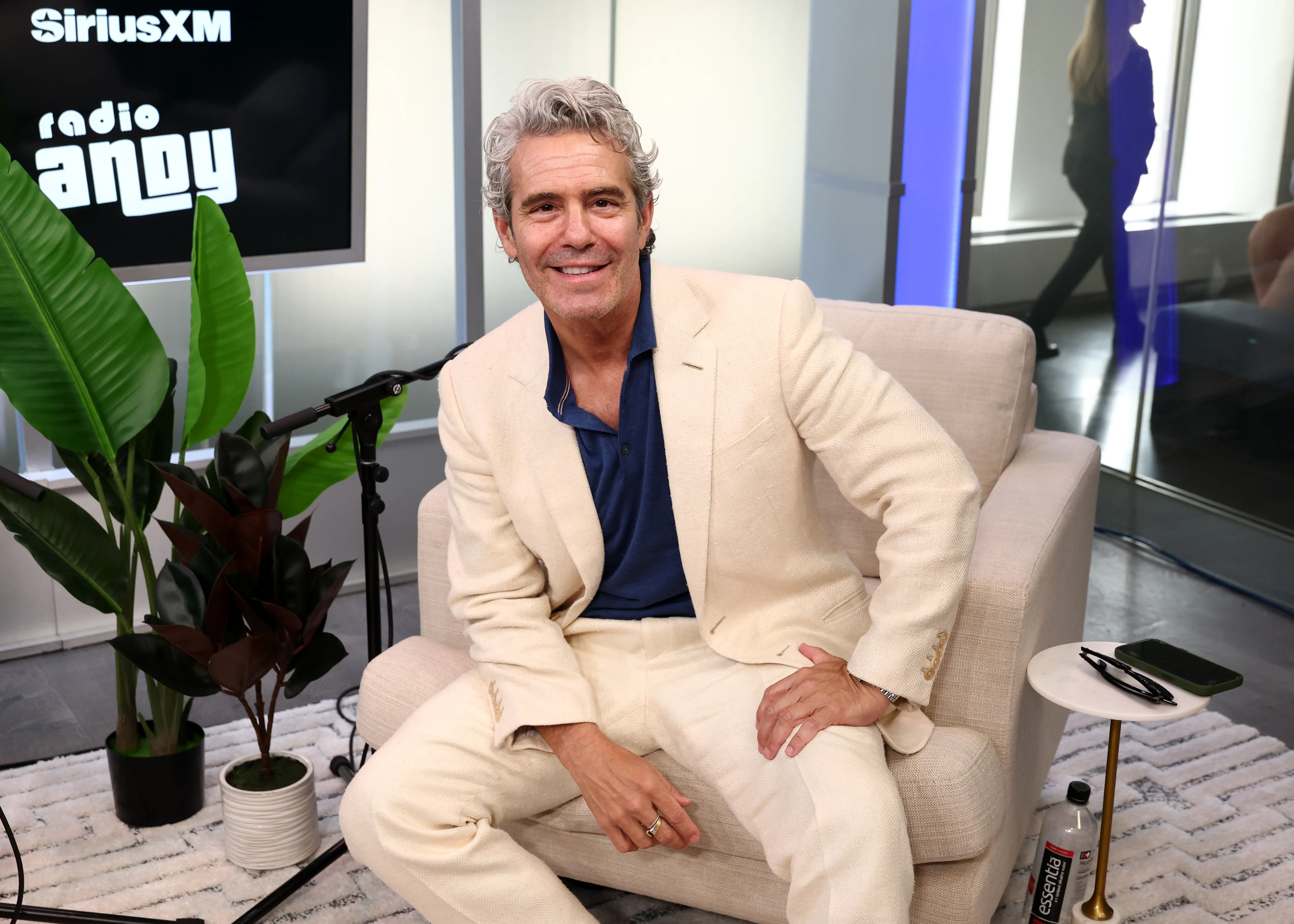 Andy Cohen smiles while sitting on a light-colored armchair with a "Radio Andy" and SiriusXM logo on a screen in the background.