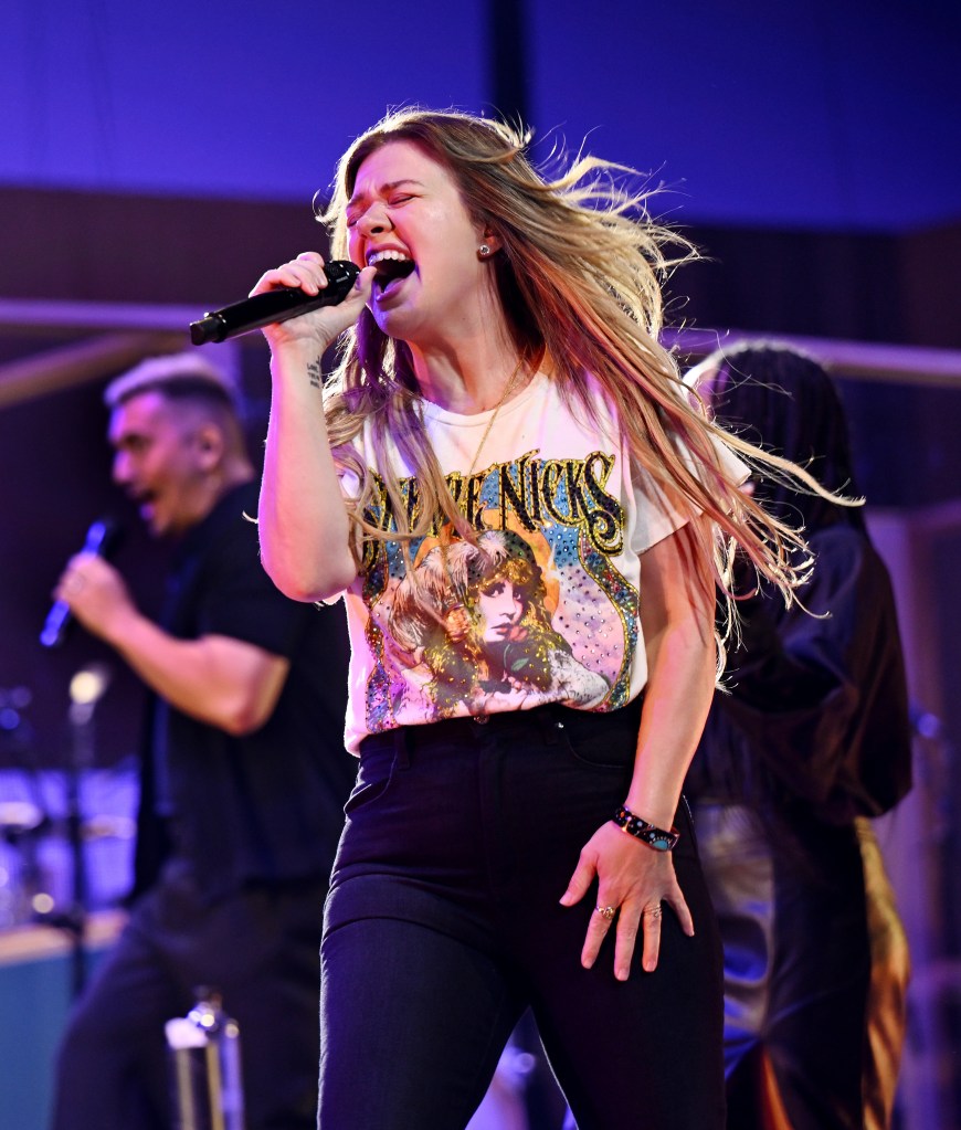 Kelly Clarkson performing during her Las Vegas residency, singing into a microphone with her mouth open and hair blowing, wearing a T-shirt and black pants.