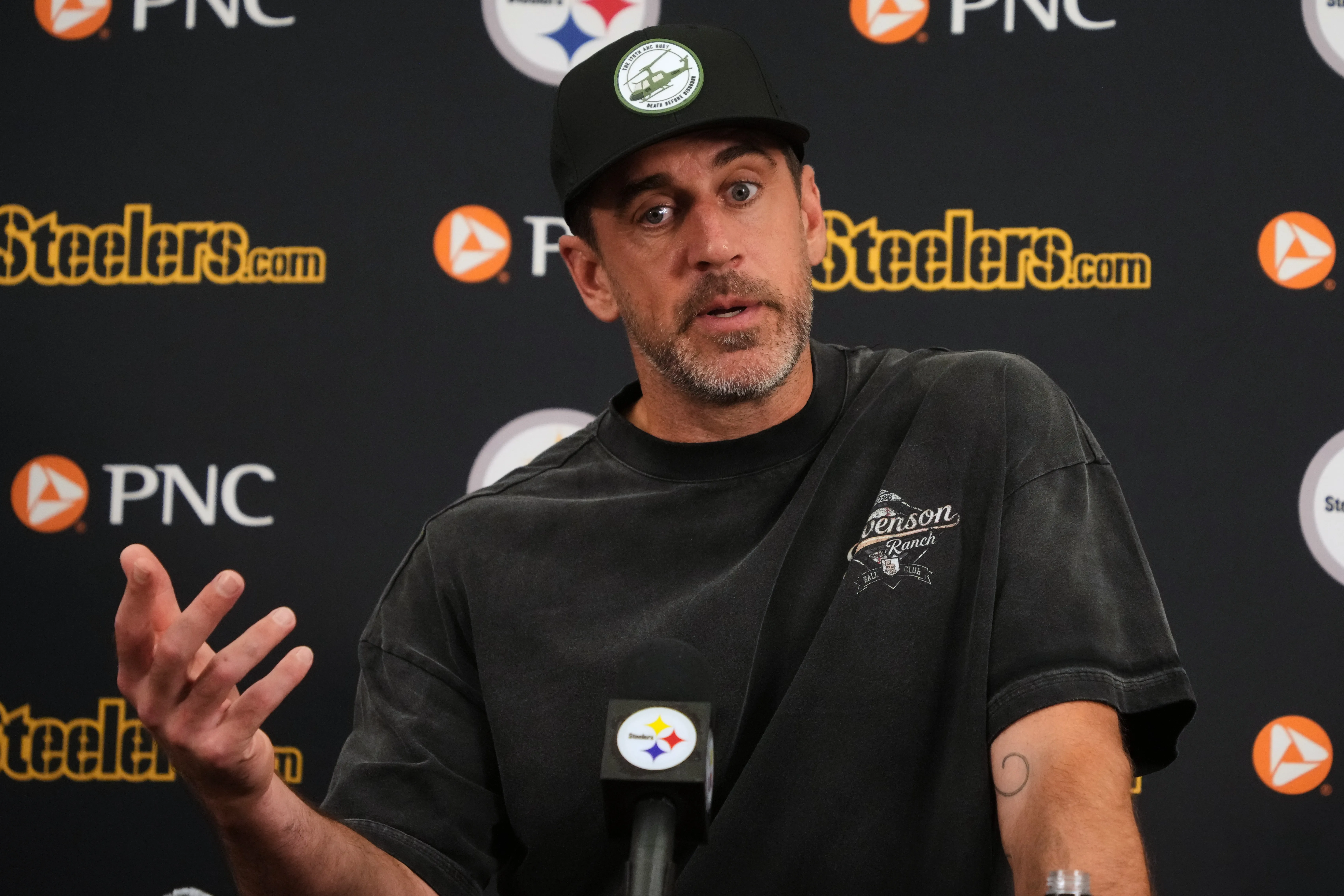 Aaron Rodgers speaking at a press conference with a Pittsburgh Steelers backdrop.