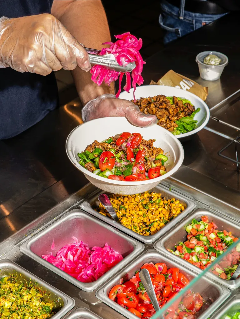 A person wearing gloves adds pickled red onions to a bowl of Mediterranean food.