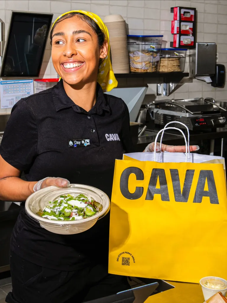 A Cava employee smiles while holding a takeout bowl and a Cava branded bag.