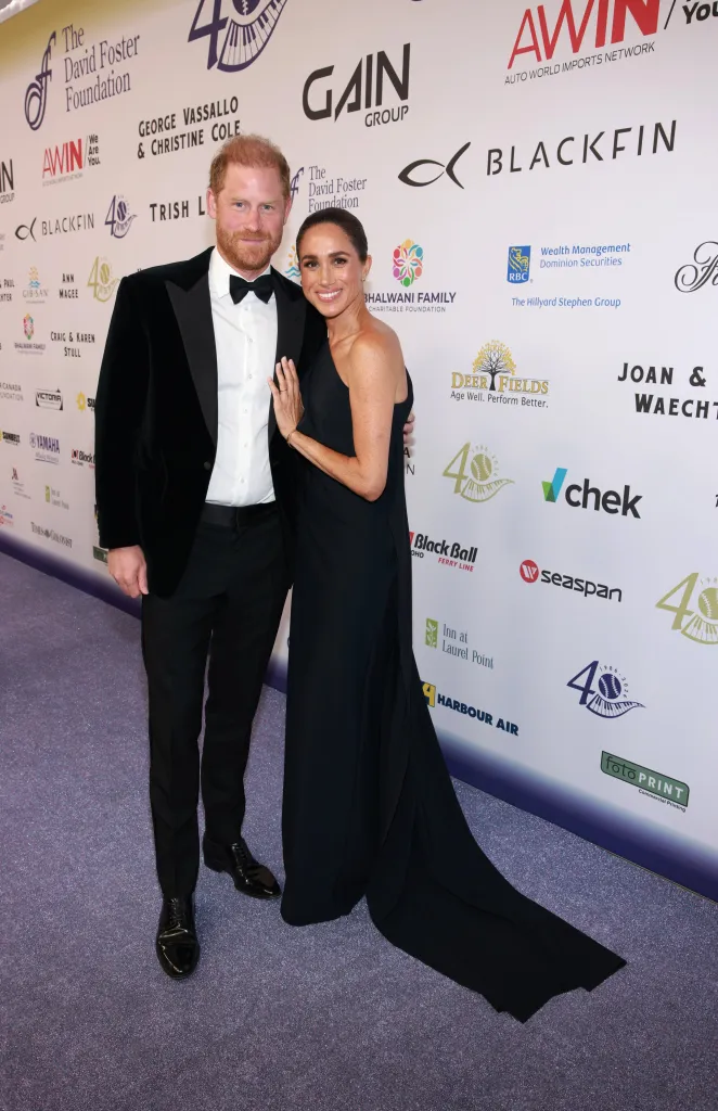 Prince Harry and Meghan, Duchess of Sussex, standing together in formal attire in front of a white backdrop with various sponsor logos.