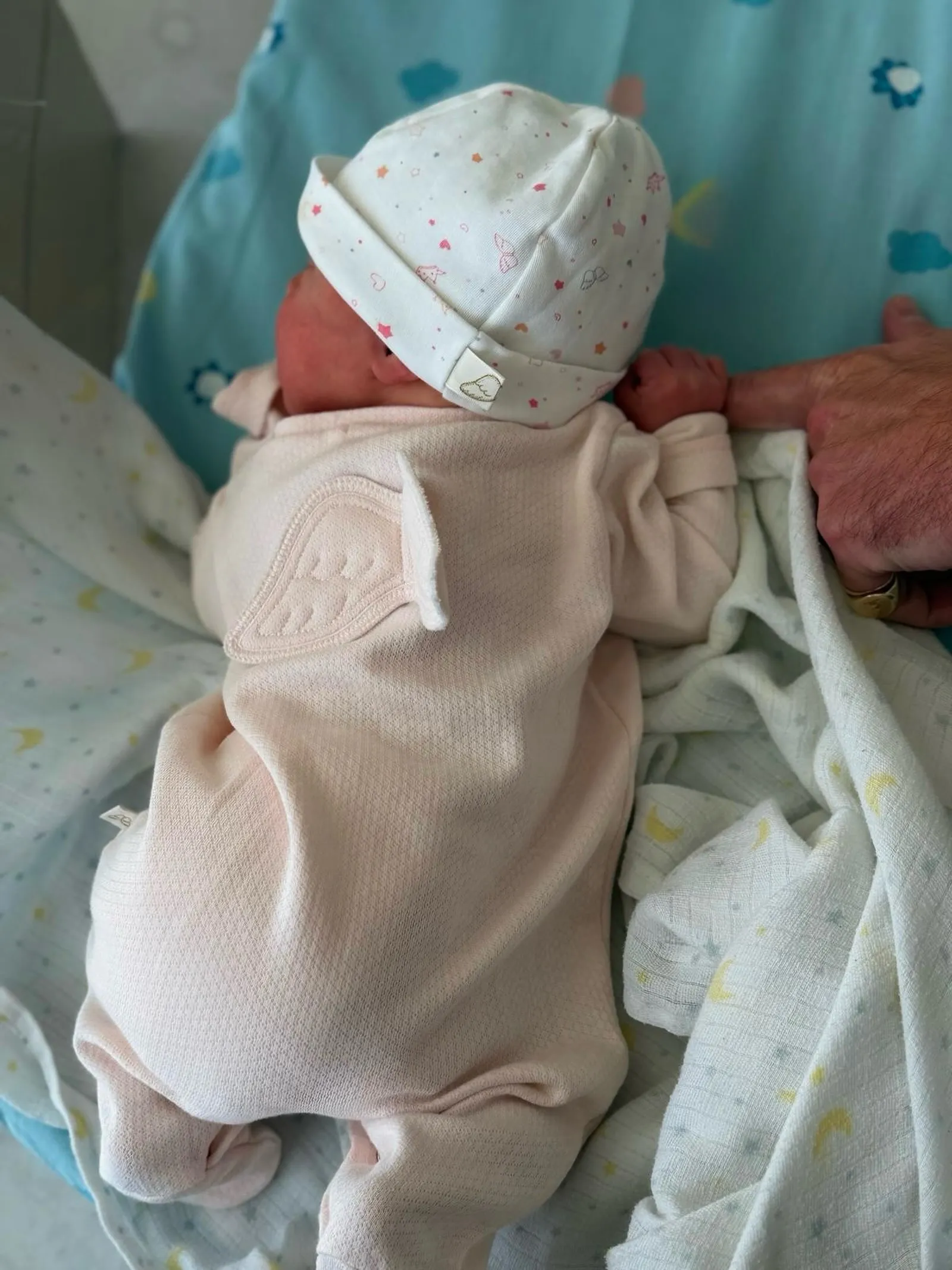 A newborn baby in a pink outfit with angel wings and a patterned hat, holding an adult's finger.