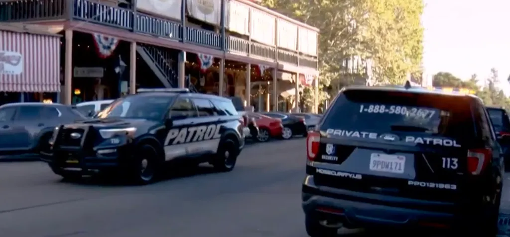 Two black private security patrol vehicles drive down a street lined with shops.