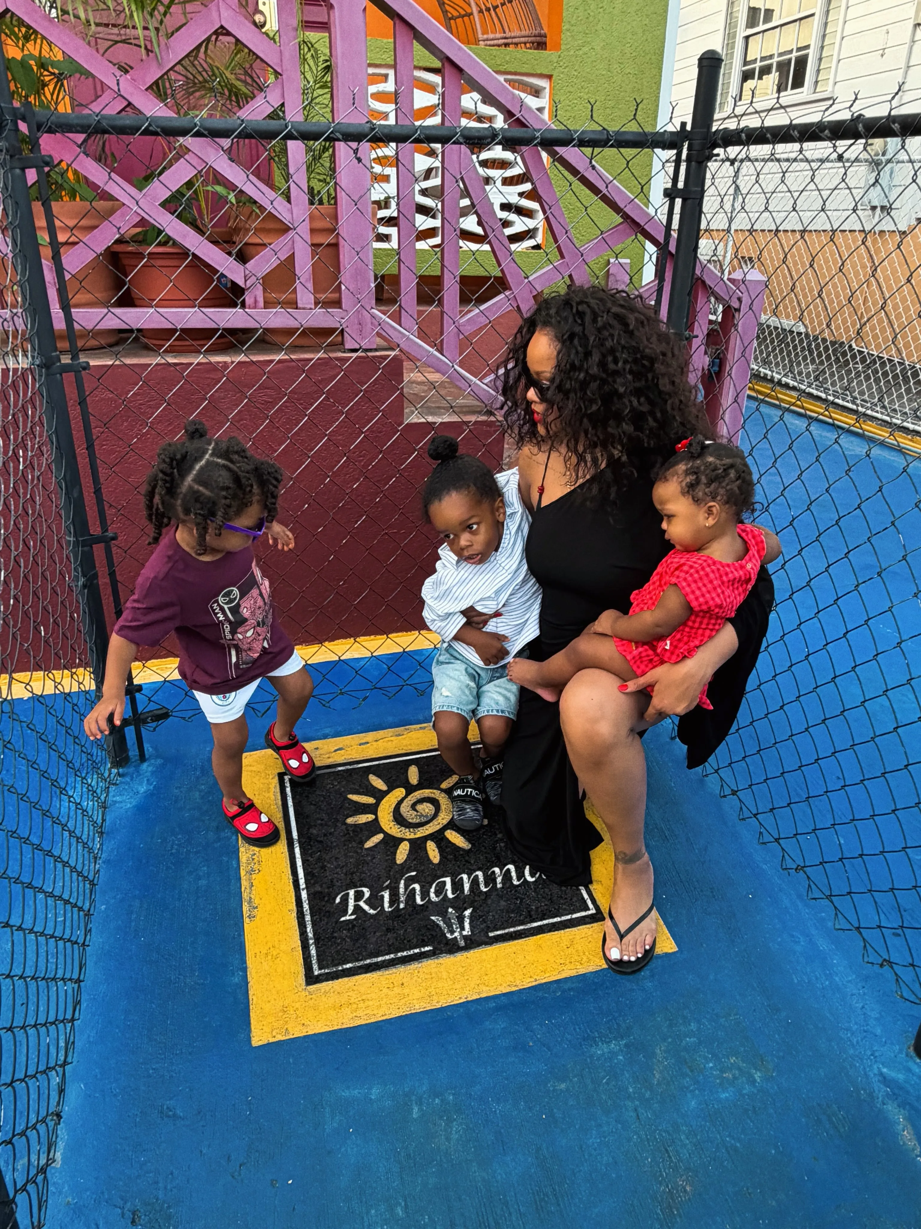 Rihanna sitting on a blue and yellow painted ground, holding her baby, while her two young sons stand nearby.