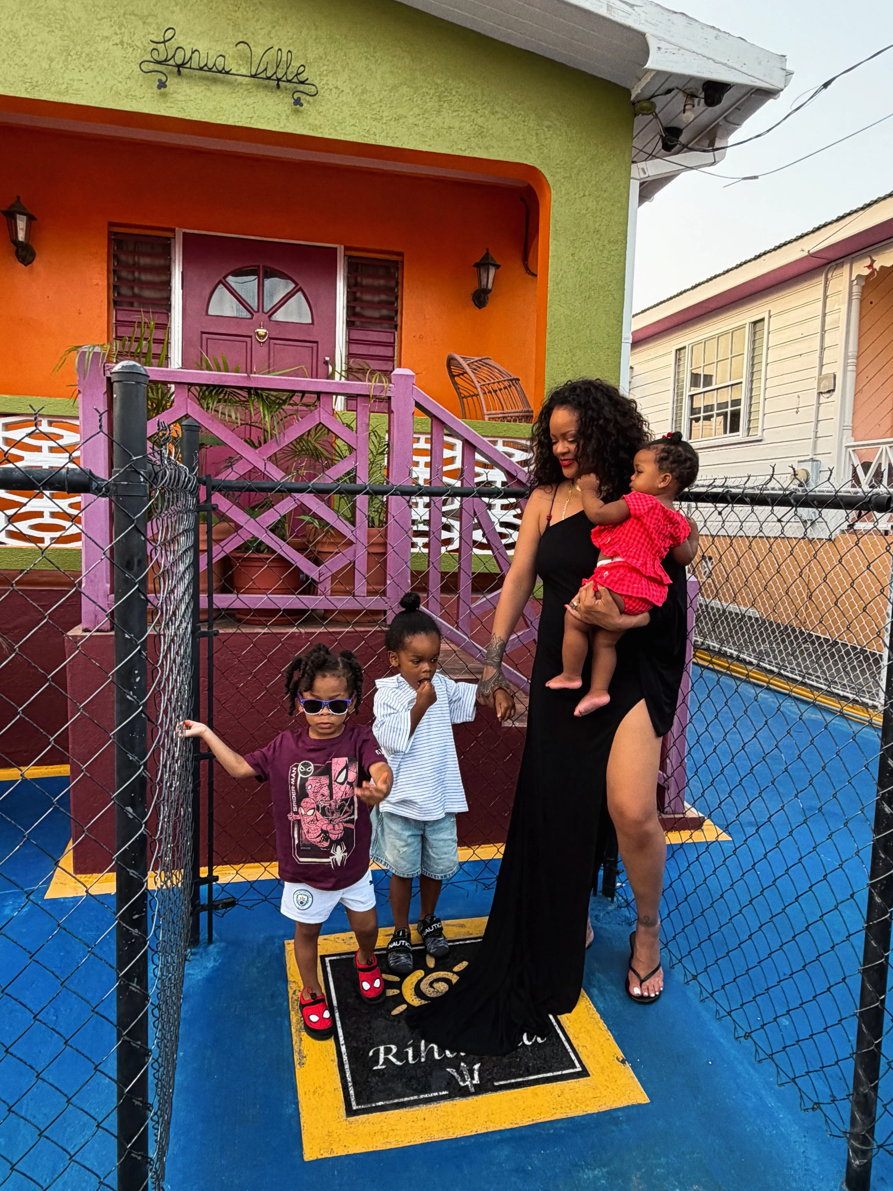 Rihanna holding her infant, with her two other children standing nearby, in front of a colorful house.