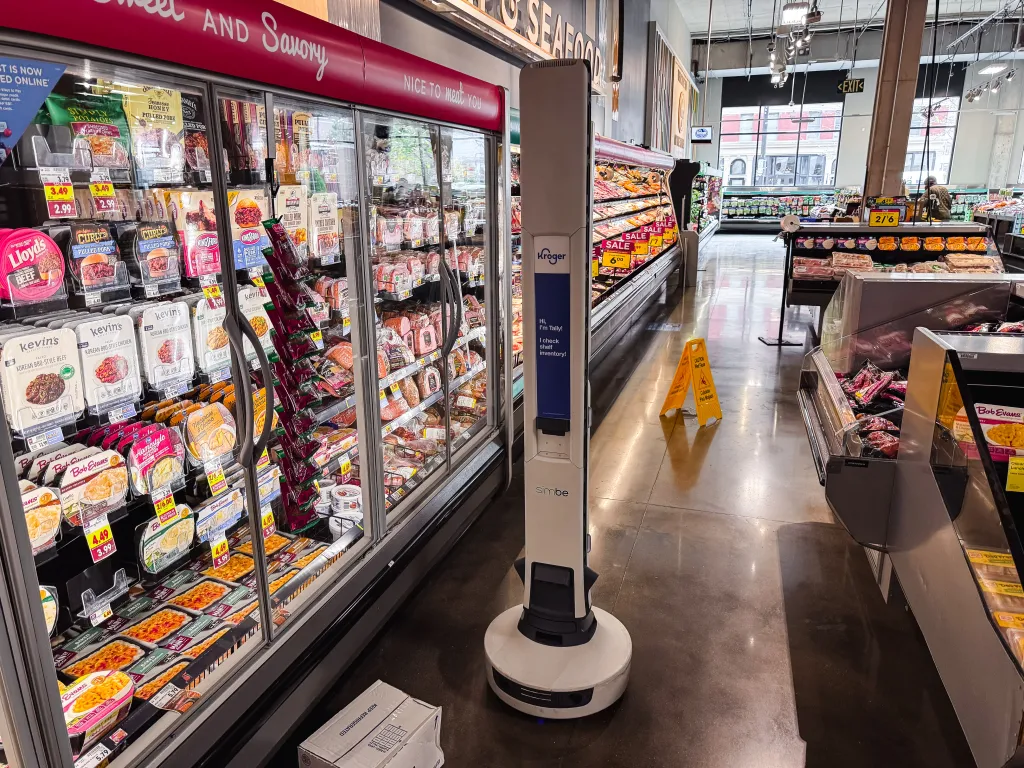 A white and blue Kroger robot named Tally scans shelves in a supermarket aisle.