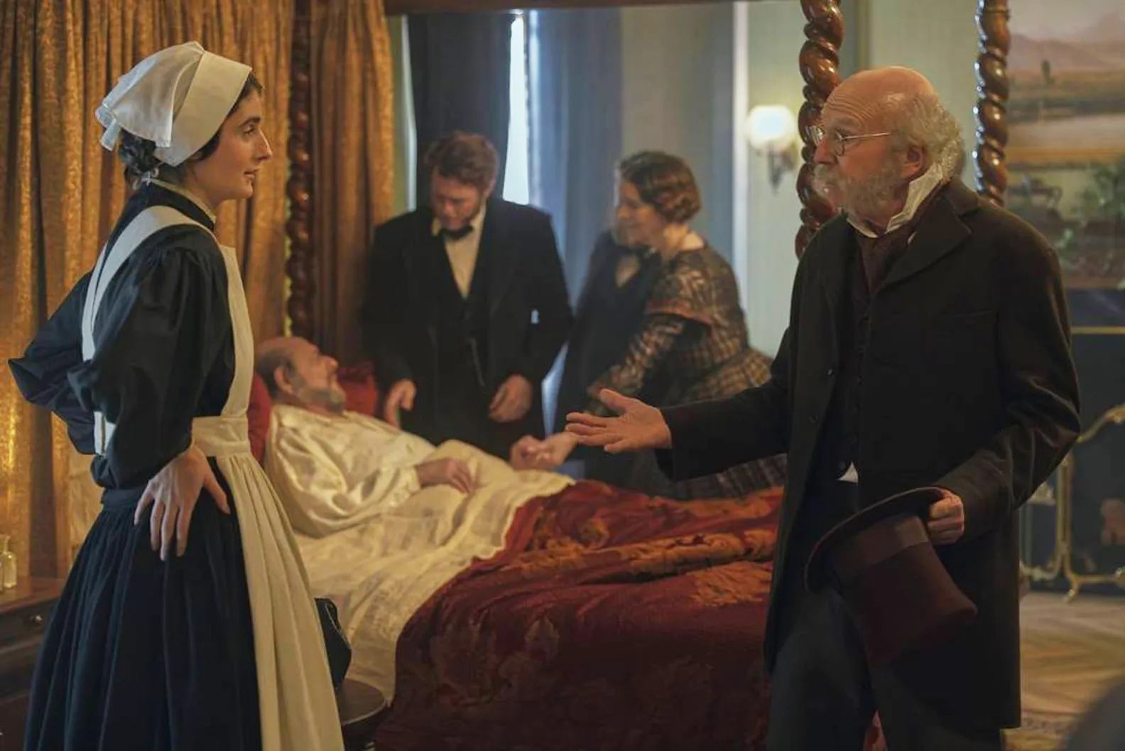 Larry David standing over a bed where another man is lying, with a woman in a maid's outfit looking on.
