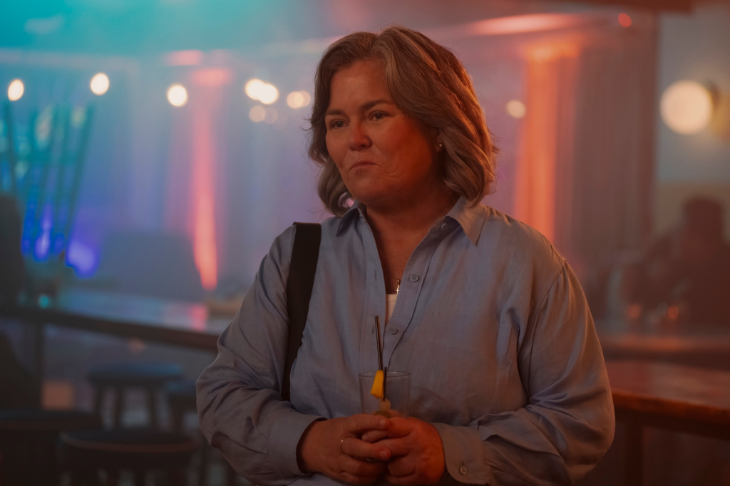 Rosie O'Donnell in a blue button-down shirt, holding a drink, with colorful lights in the background.
