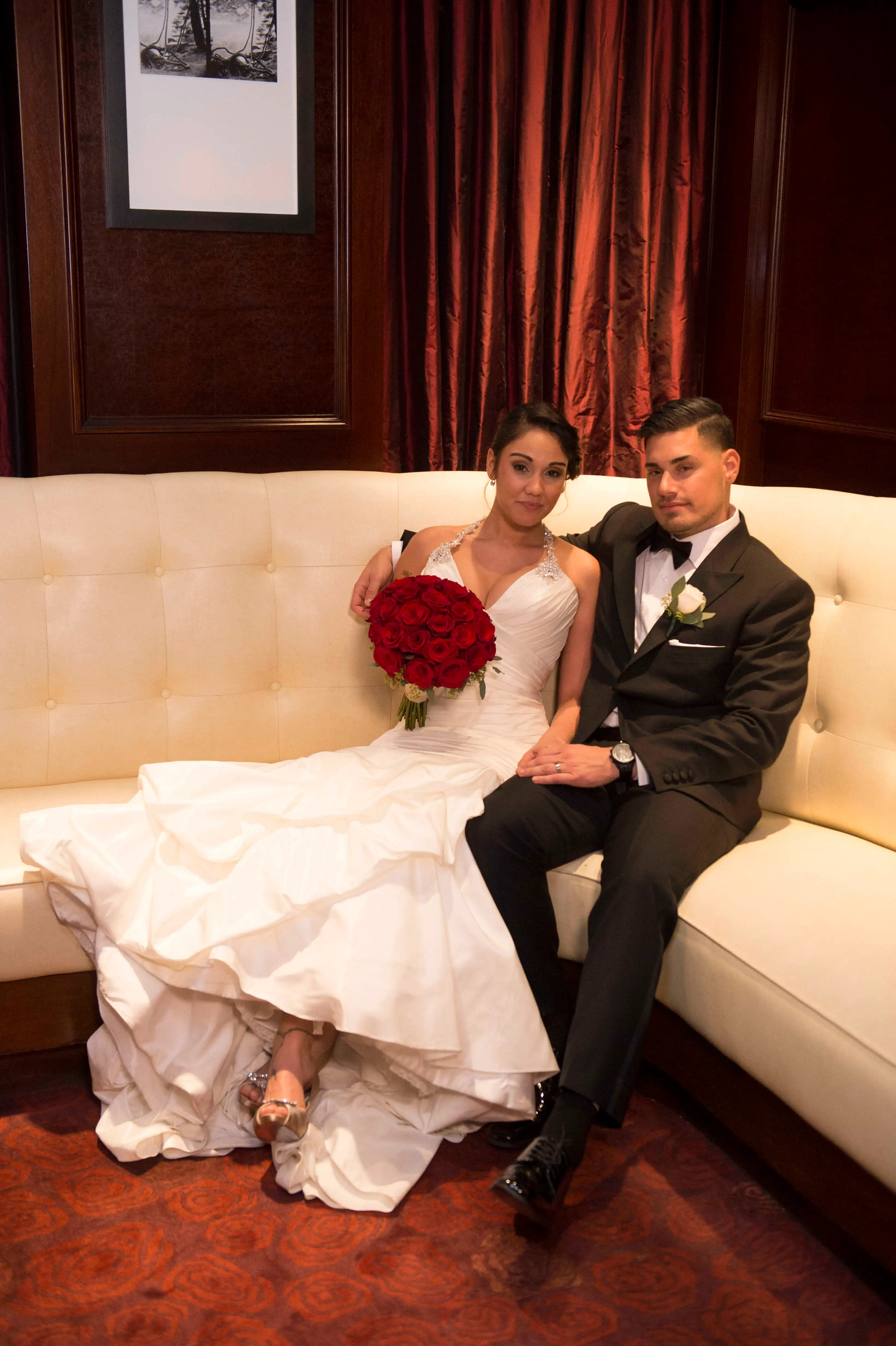 Ryan De Nino and Jessica Castro in wedding attire sitting on a cream-colored couch.