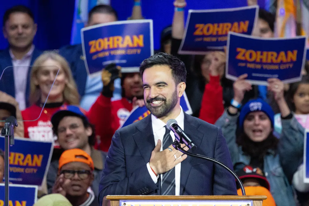 Zohran Mamdani smiling at a rally, with supporters holding "Zohran for New York City" signs in the background.