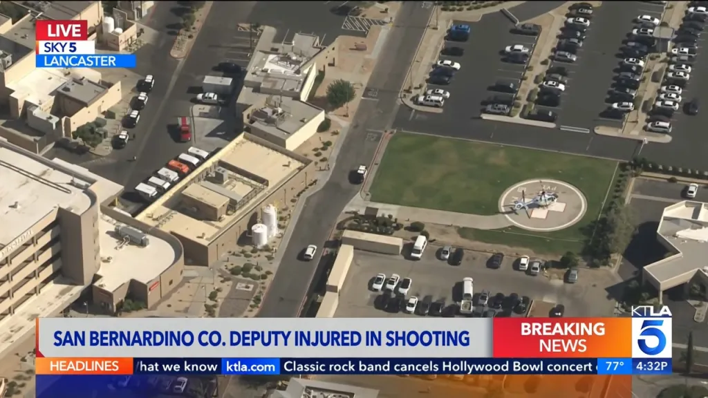 Aerial view of a hospital with a helicopter on a helipad and several emergency vehicles, with a news chyron at the bottom reading "SAN BERNARDINO CO. DEPUTY INJURED IN SHOOTING."