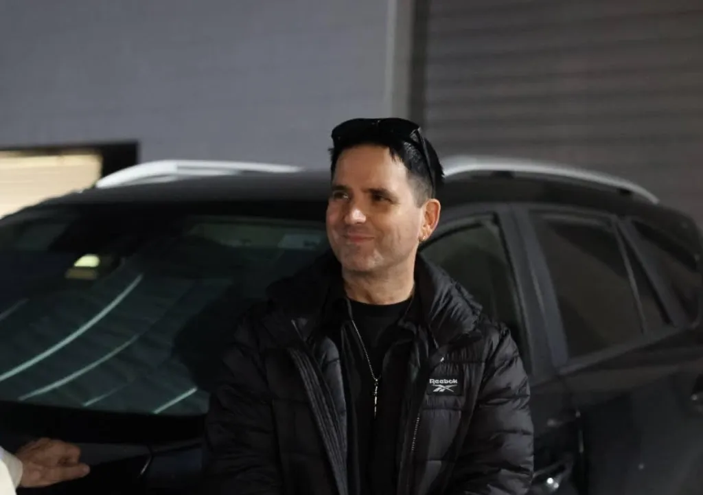 Shane Ryan-Reid, wearing a black puffer jacket and sunglasses on his head, smiles while standing in front of a dark car.