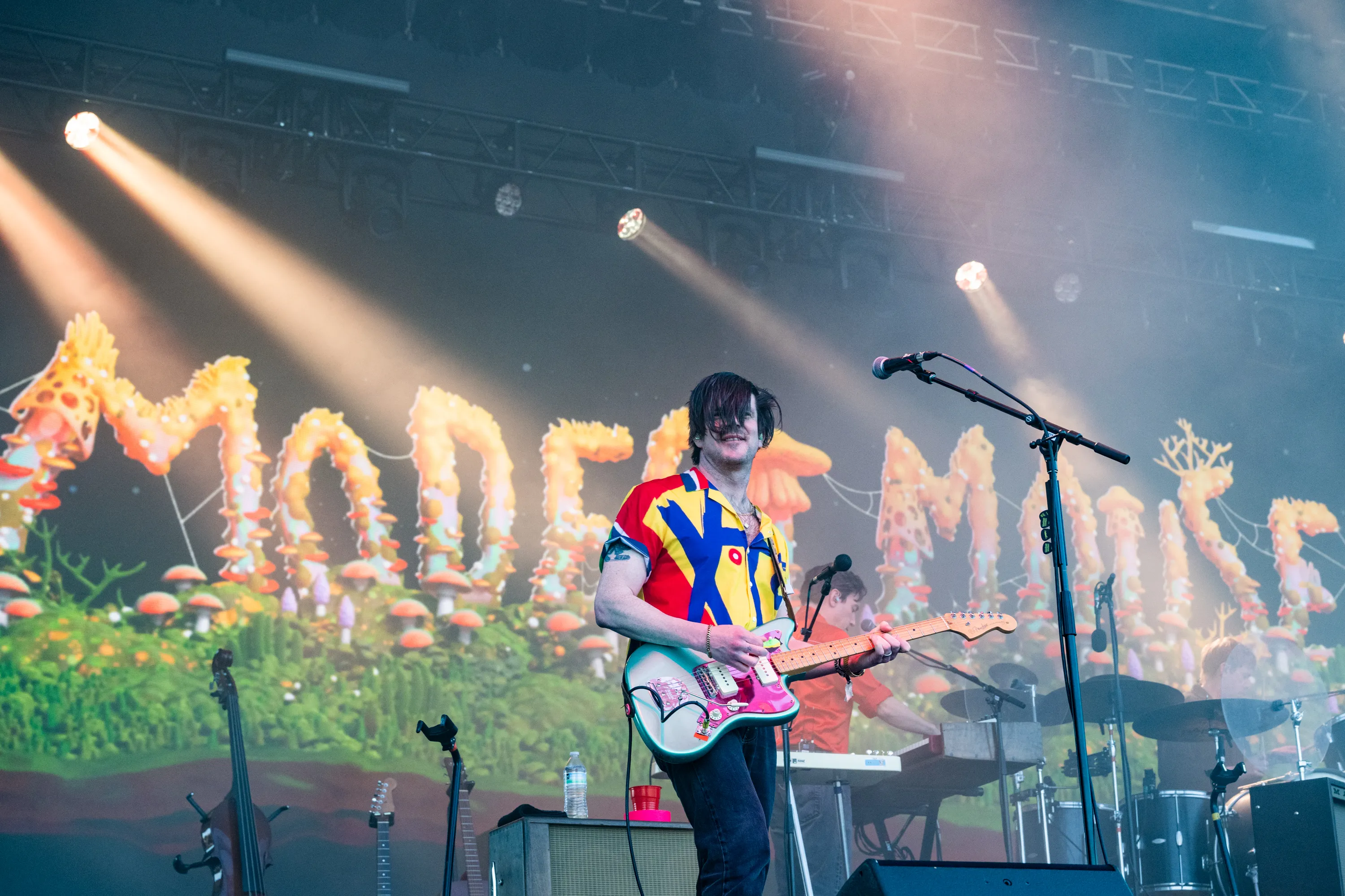 Simon O'Connor of Modest Mouse performs live on stage, playing a light blue electric guitar with pink accents, wearing a red, yellow, and blue patterned shirt.