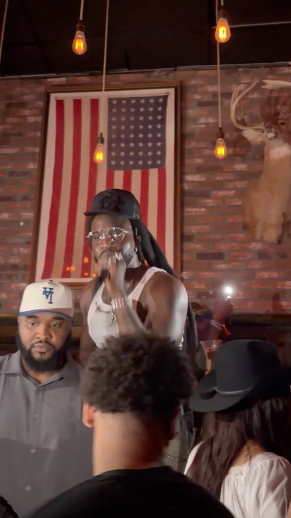 Shaboozey performs while wearing a white tank top, a black baseball cap, and glasses, with an American flag and deer head mounted on the brick wall behind him.