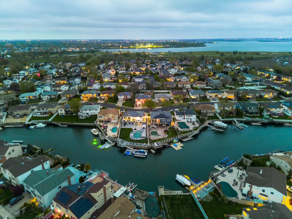 An aerial panoramic view shows a small illuminated town on sea channel bank with boats and yachts on piers against cloudy sky, twilight in Nassau County, New York.