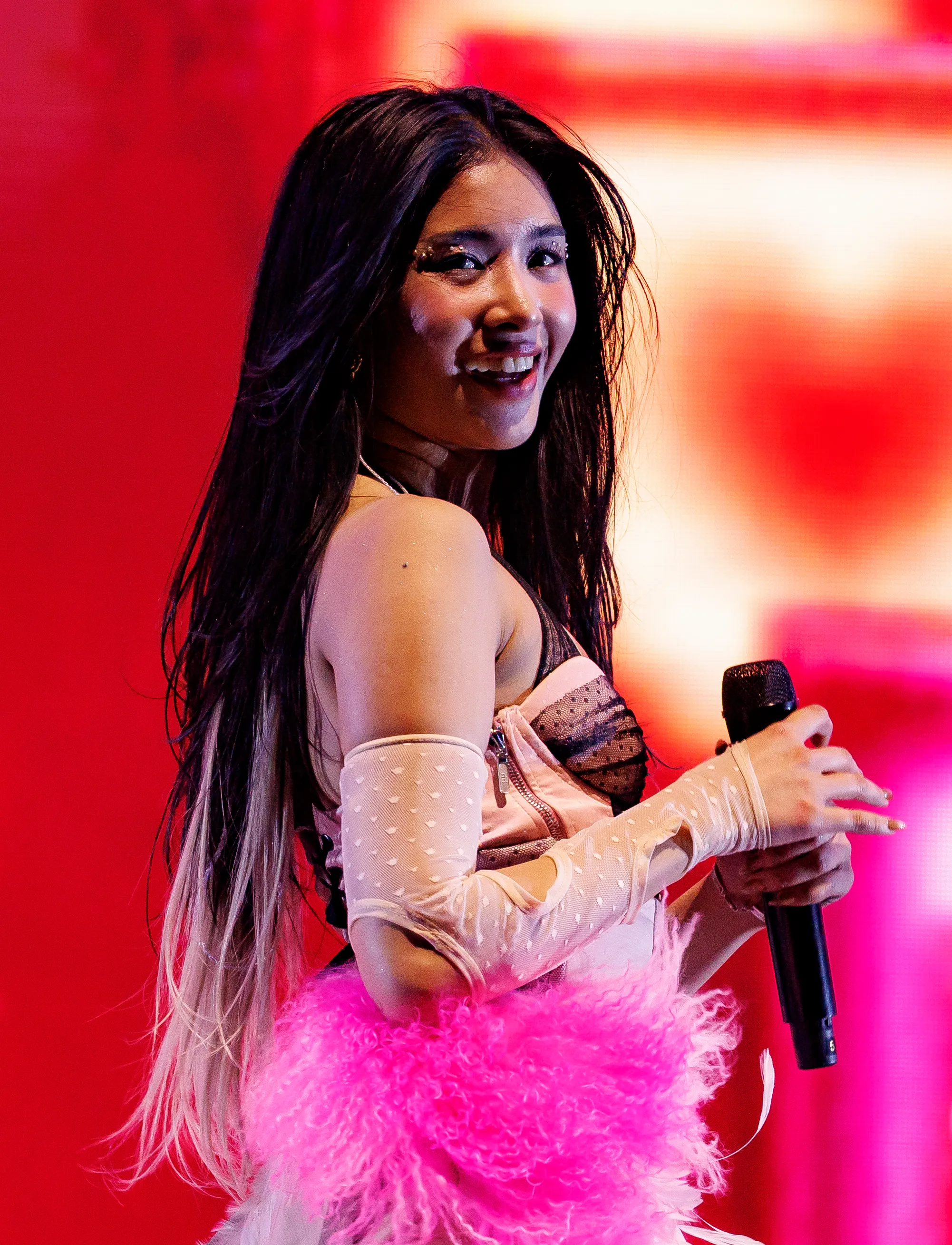 Sophia Laforteza of KATSEYE performing with a microphone, wearing a pink outfit with feathers and long gloves.