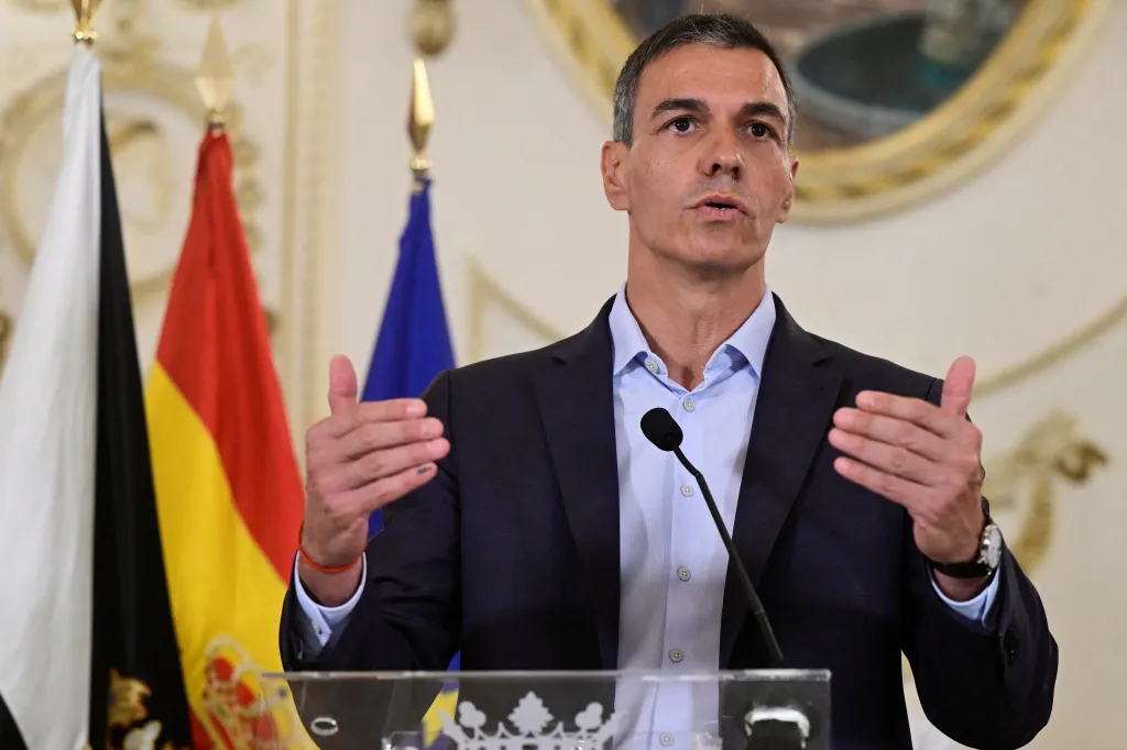Spanish Prime Minister Pedro Sanchez speaks at a press conference, gesturing with his hands, with flags behind him.