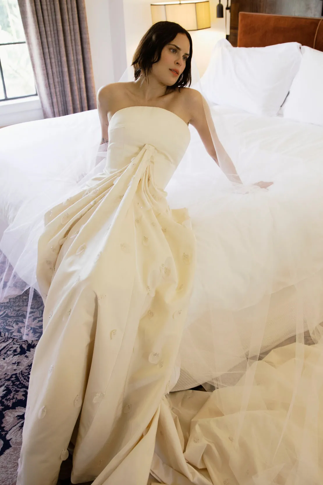 Tallulah Willis in a strapless silk satin Balenciaga wedding gown with floral embellishments, sitting on a bed.