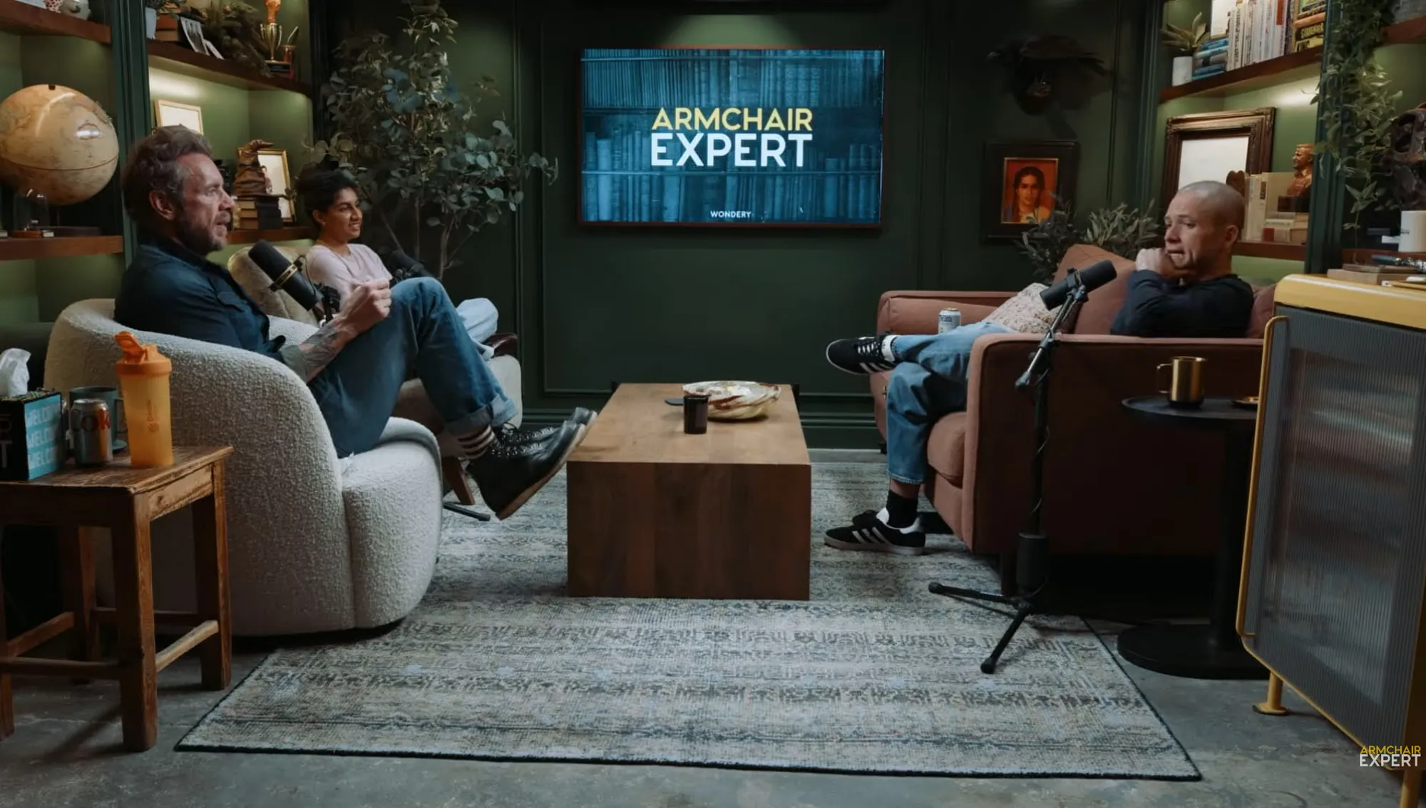 Three people sitting in armchairs, two men and one woman, conversing in a podcast studio with a screen displaying "Armchair Expert" in the background.