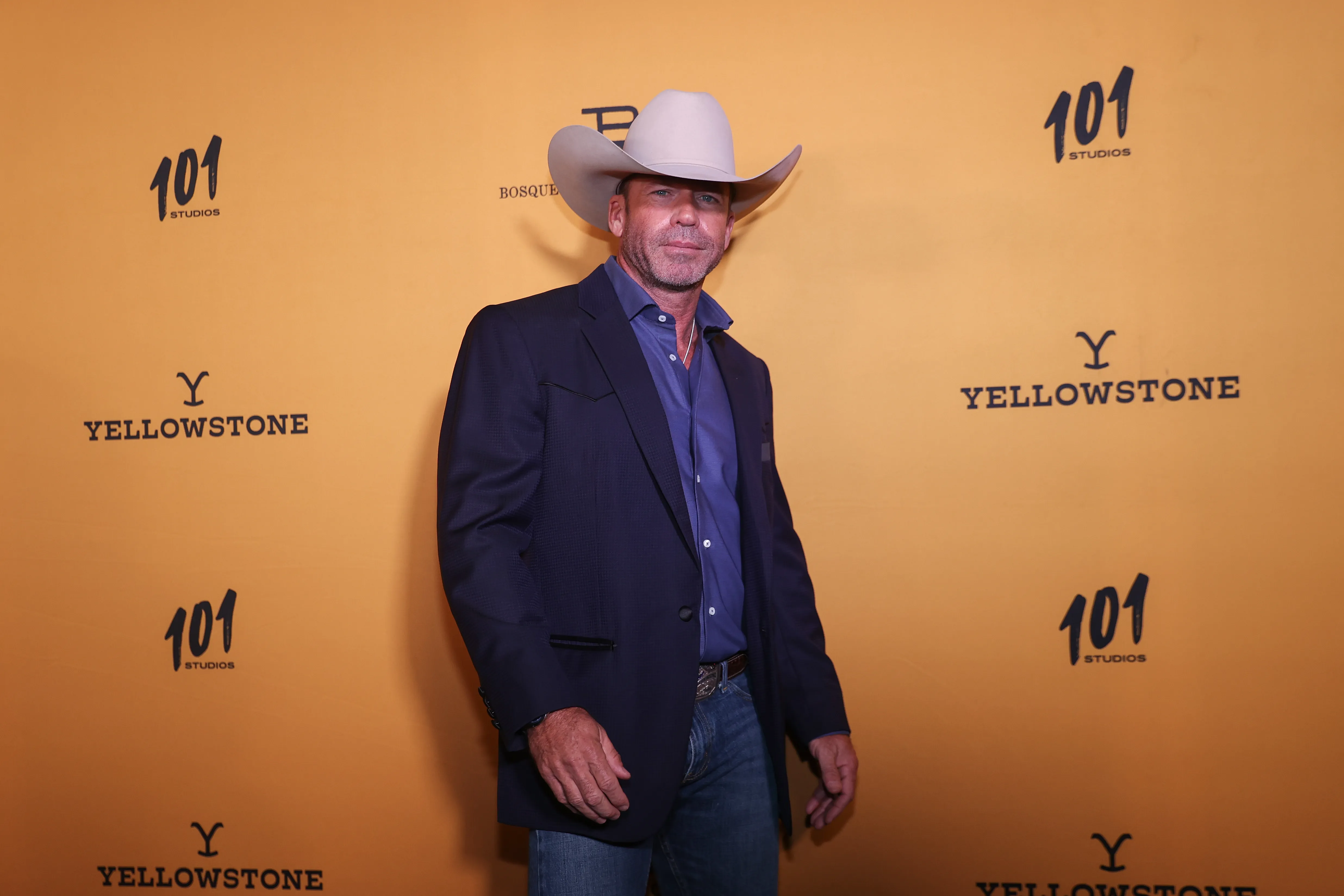 Taylor Sheridan in a cowboy hat and blue suit jacket against a yellow "Yellowstone" backdrop.