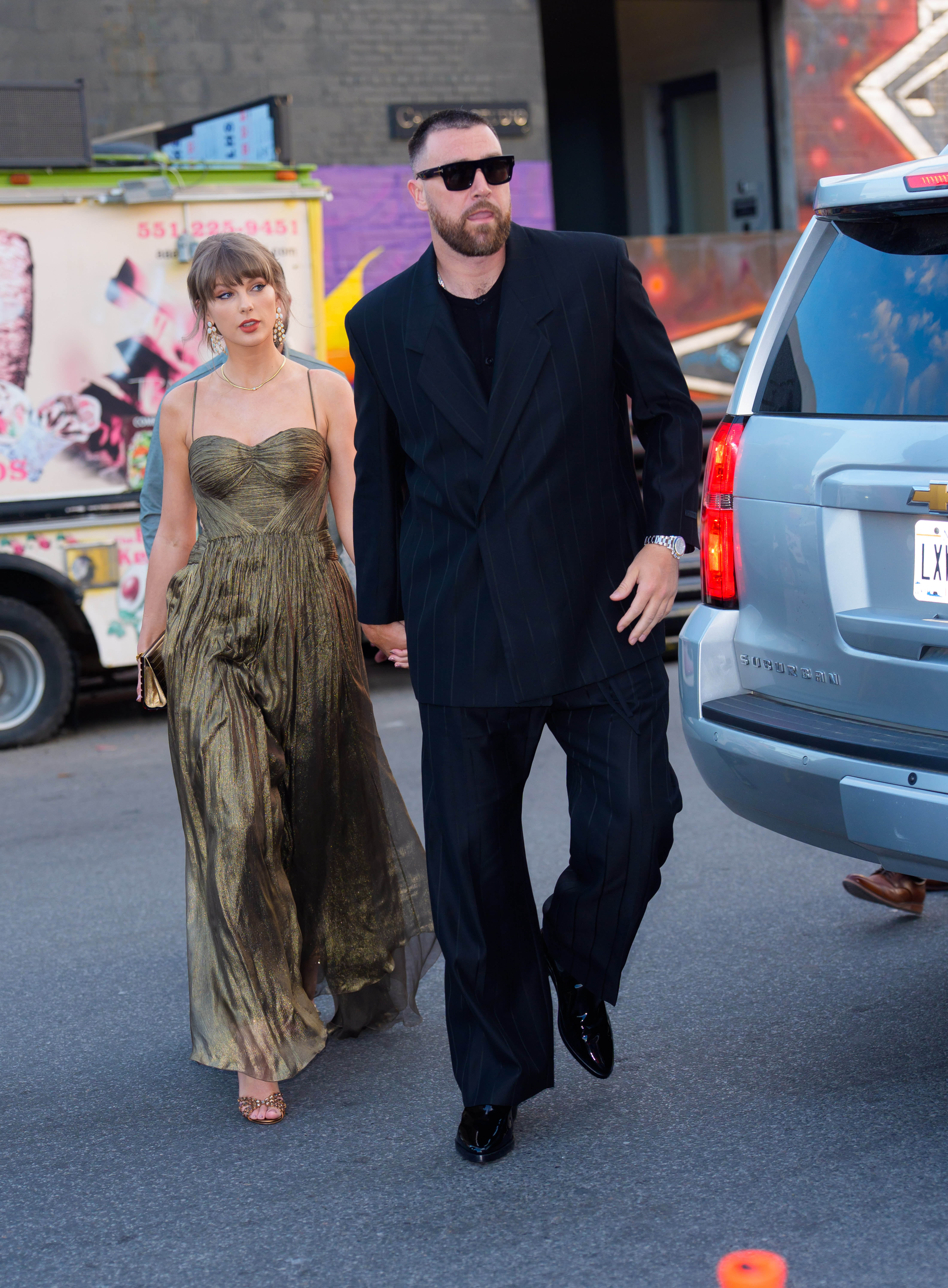 Taylor Swift in a gold dress holding hands with Travis Kelce in a dark suit.