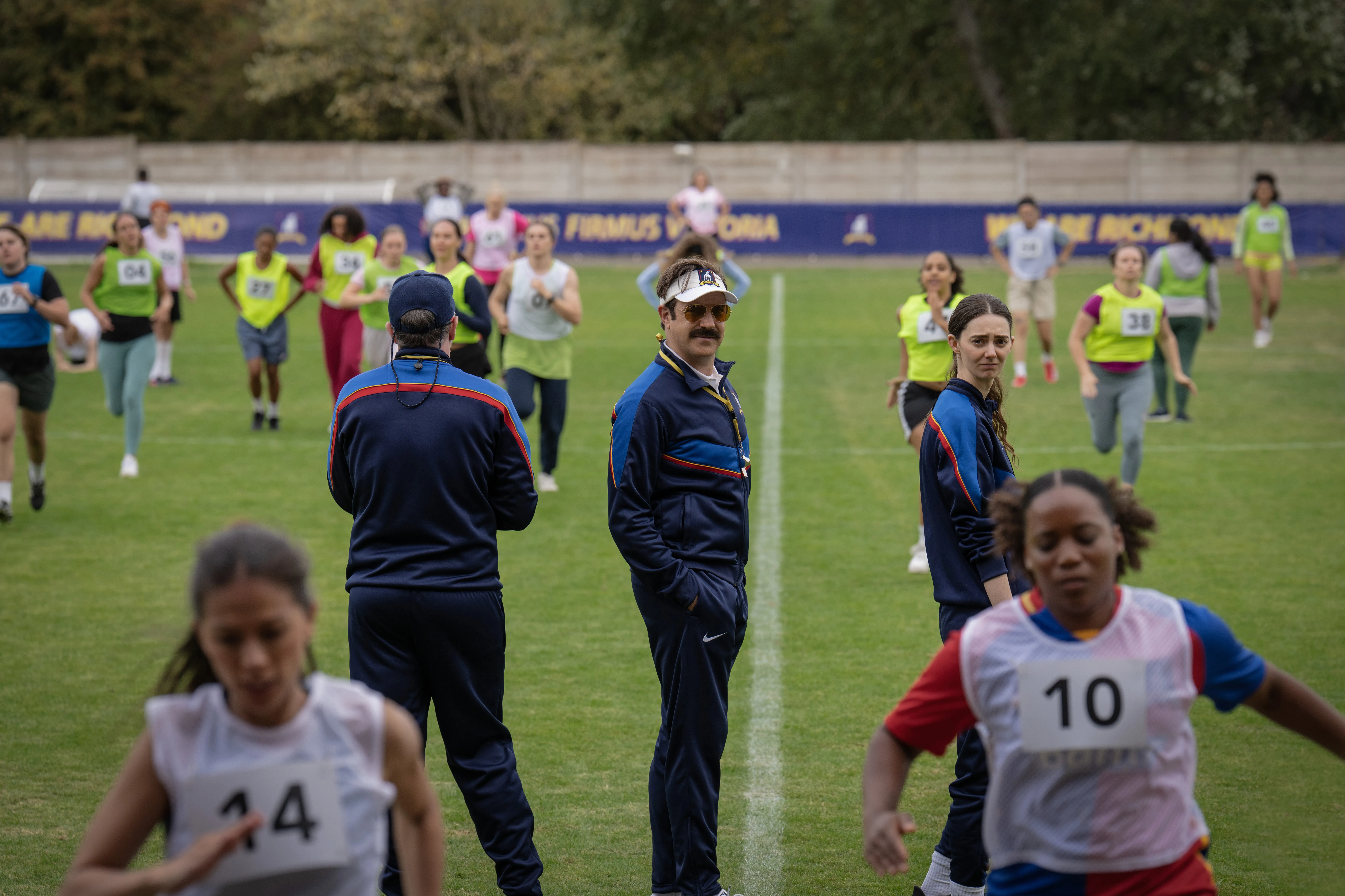 Ted Lasso and Coach Beard watching women's soccer players in numbered vests run drills on a field.