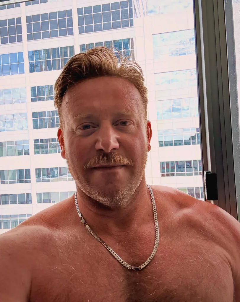 Tyler Duckworth, shirtless, looking at the camera with a city building in the background