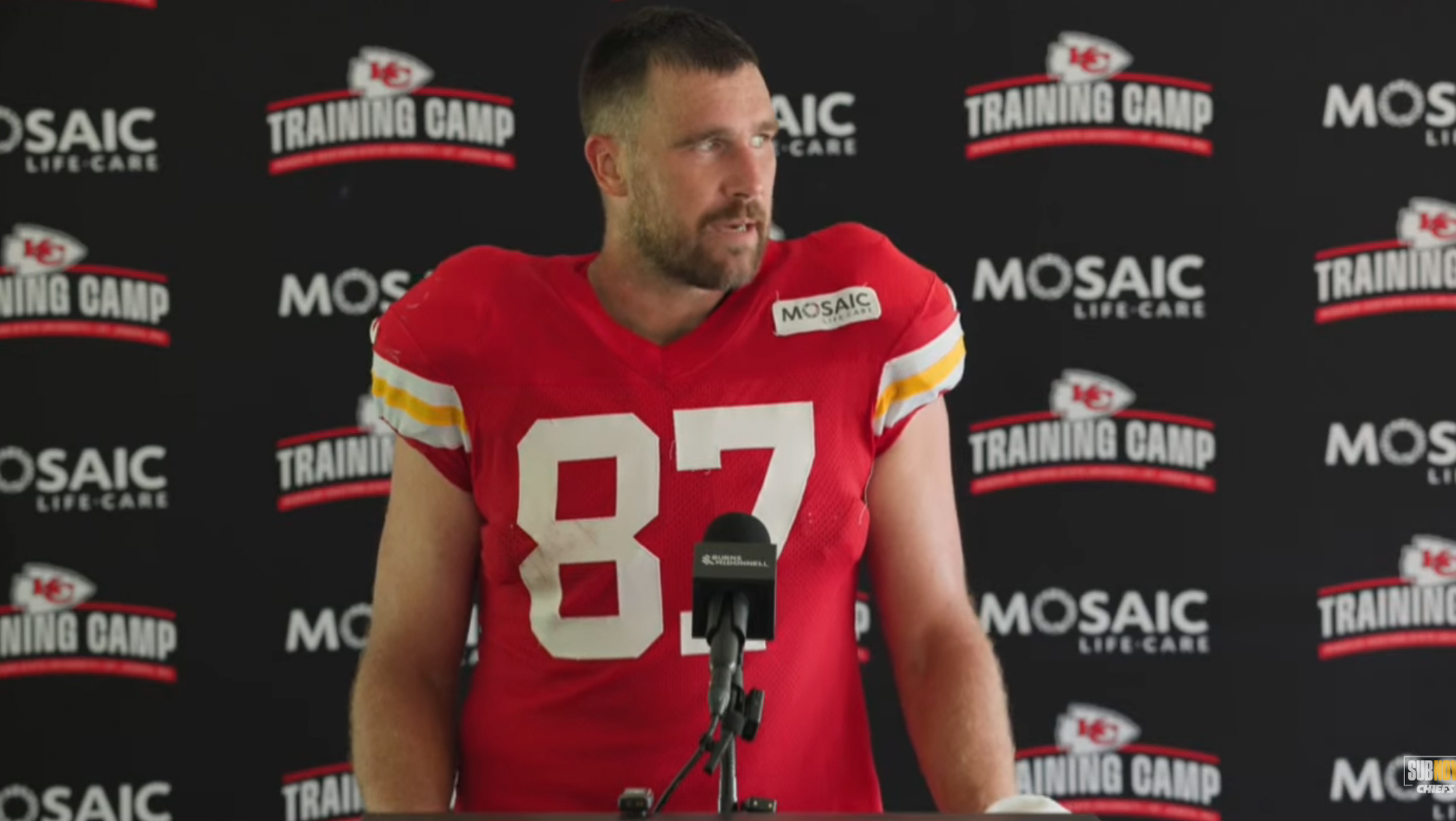 Travis Kelce in a red Kansas City Chiefs jersey with the number 87, speaking at a press conference during training camp.