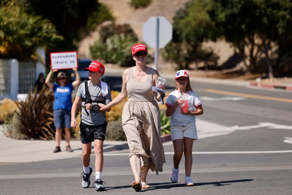 Trump supporters, including a mother and two children, walk with a woman holding a "Nuremberg Trials 2.0" sign in the background.