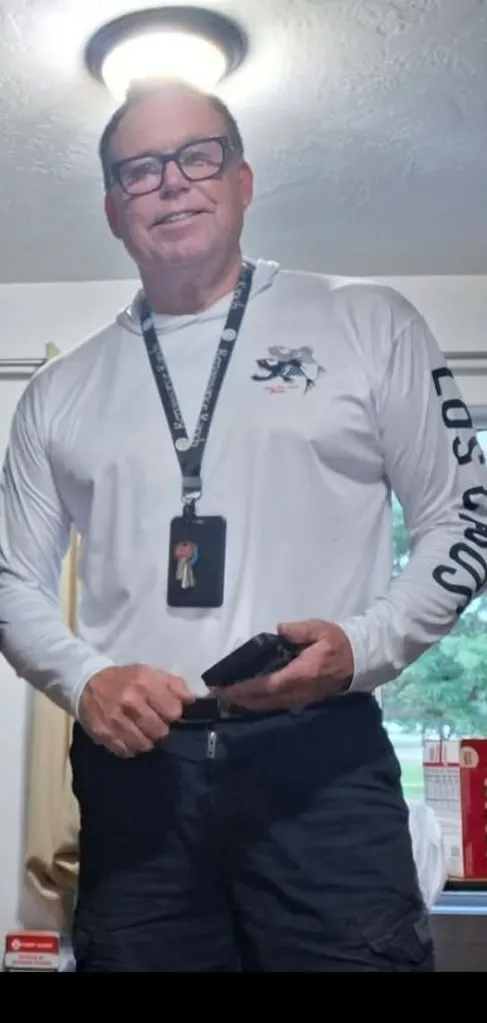 Christopher Claunch, wearing glasses and a long-sleeved shirt with a lanyard, stands indoors, smiling faintly.