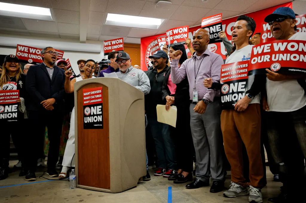 Uber and Lyft drivers celebrating at a press conference, holding signs that say "We Won Our Union".