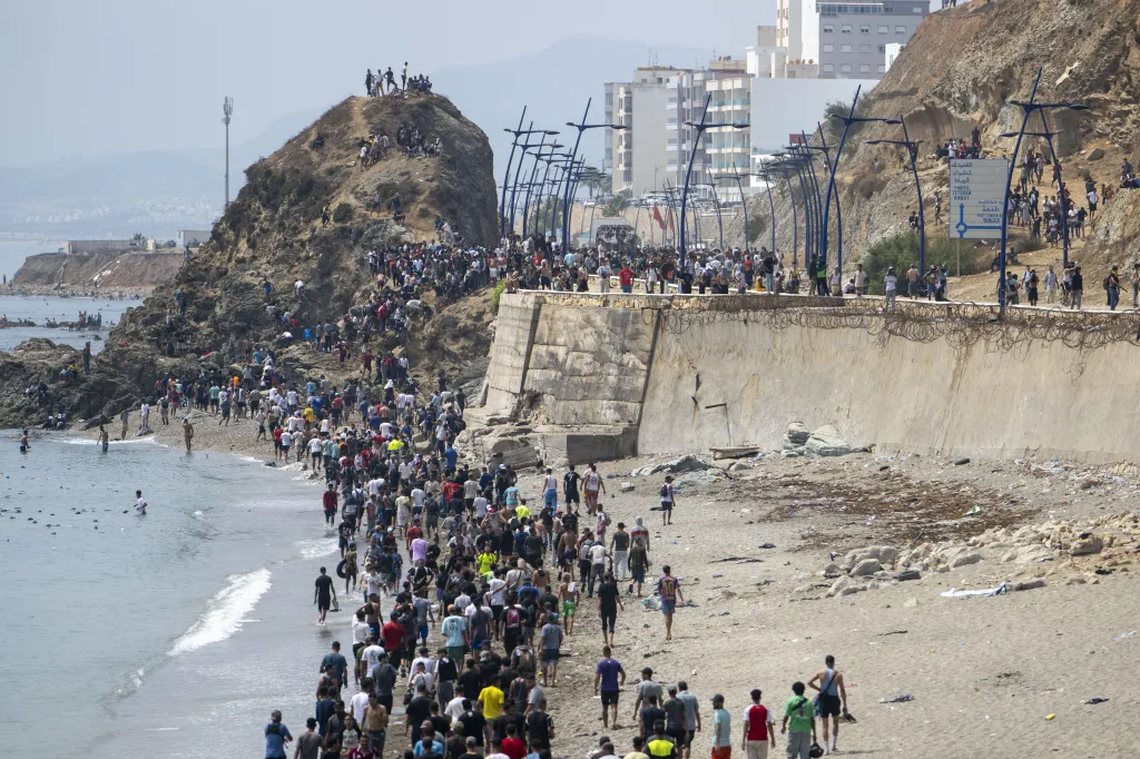 Migrants, mostly young men, walk along a beach and rocky hillside near the Spanish enclave of Ceuta, with some in the water and others climbing up cliffs.