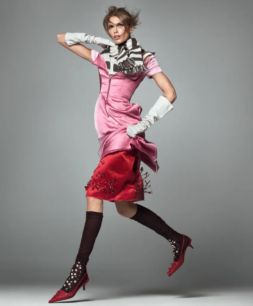 A woman in a pink dress, red skirt, and red heels with brown leg coverings and a striped scarf jumps in the air.