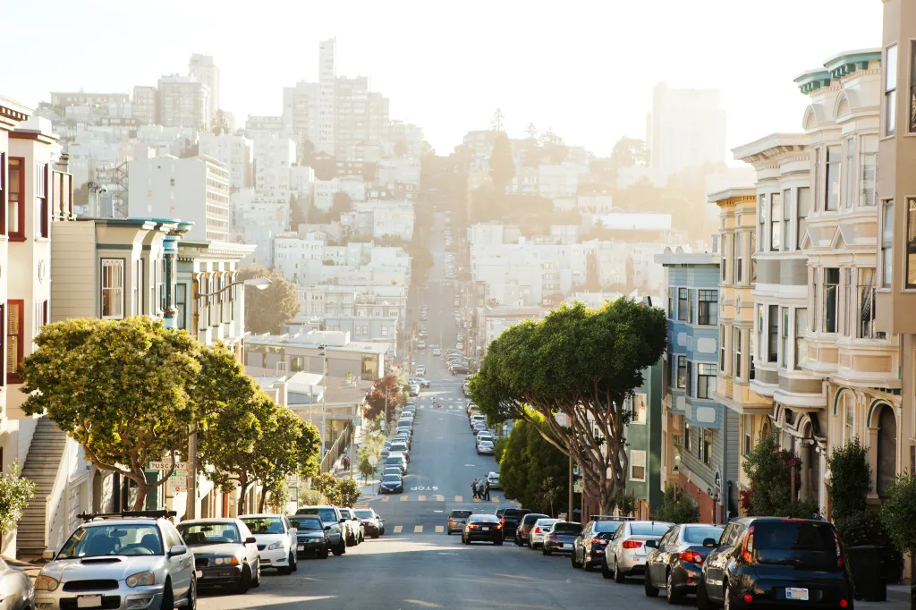 A San Francisco street descends downhill, lined with parked cars and multi-story homes, with tall buildings in the background.