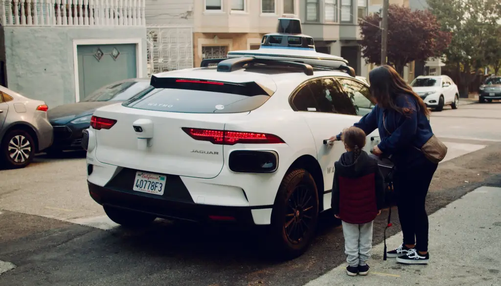 A person and child standing next to a white Waymo self-driving car.