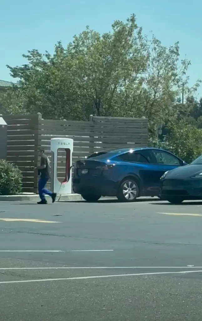 A person runs with a rifle past a Tesla charging station and two parked cars.