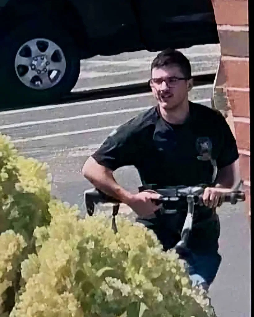 A man in glasses holding a long gun partially obscured by bushes.