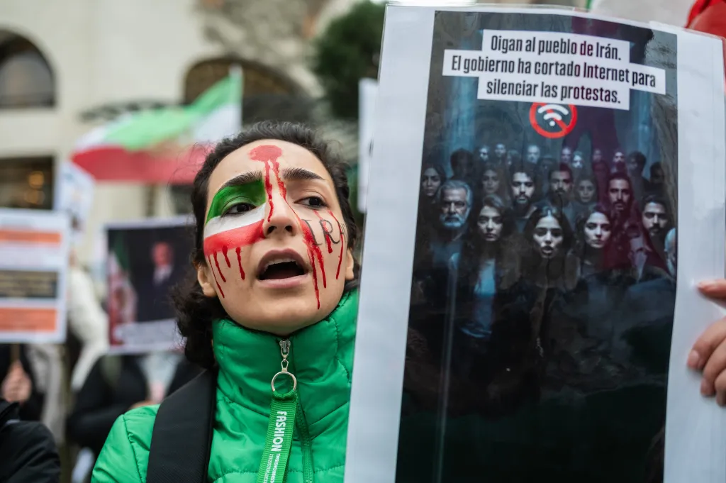 A woman with her face painted like the Iranian flag and red streaks resembling blood, holds a sign that reads, "Listen to the people of Iran. The government has cut off the Internet to silence protests."