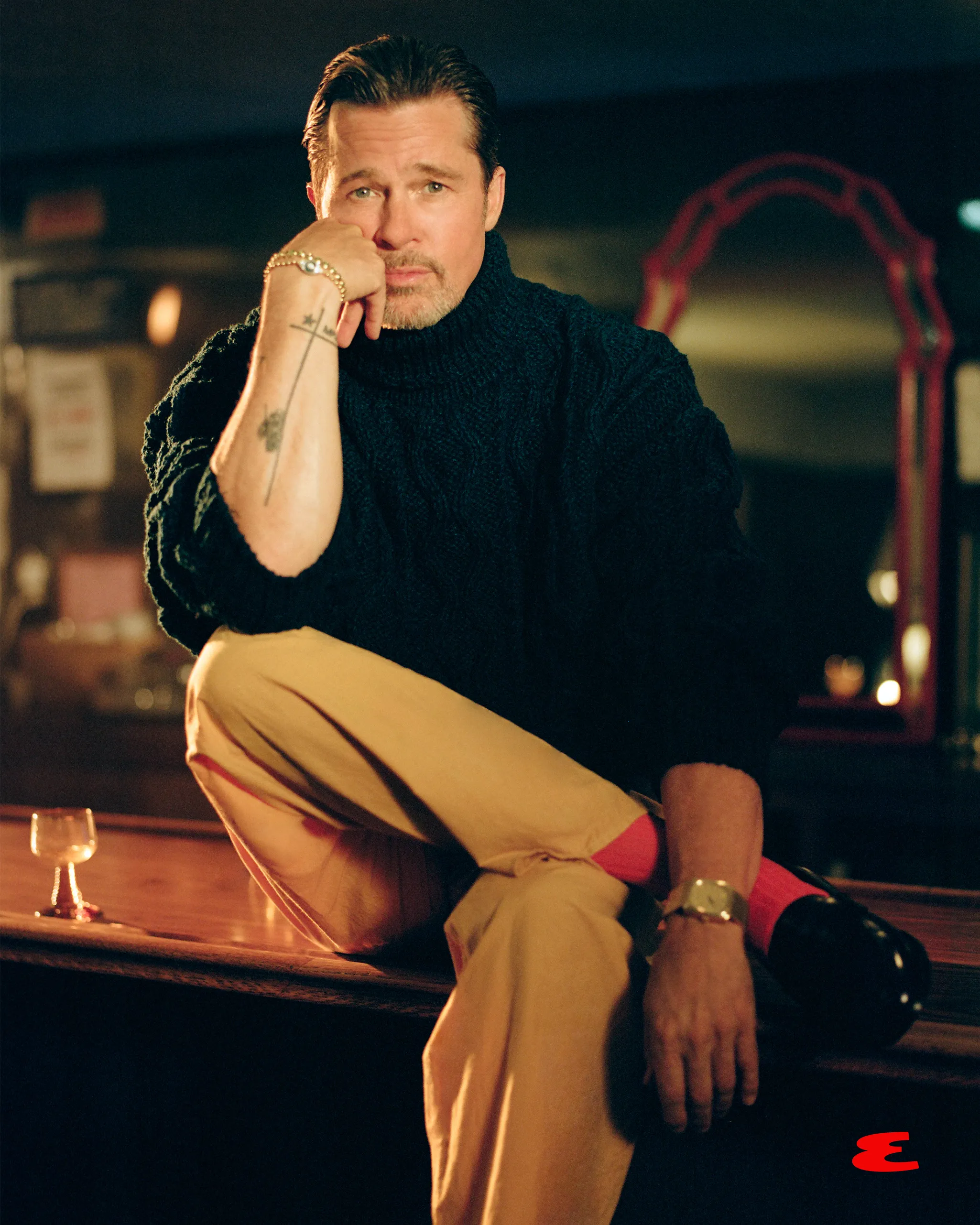 Brad Pitt wearing a dark green turtleneck and light brown pants, sitting on a bar, with his elbow on his knee and chin in his hand.