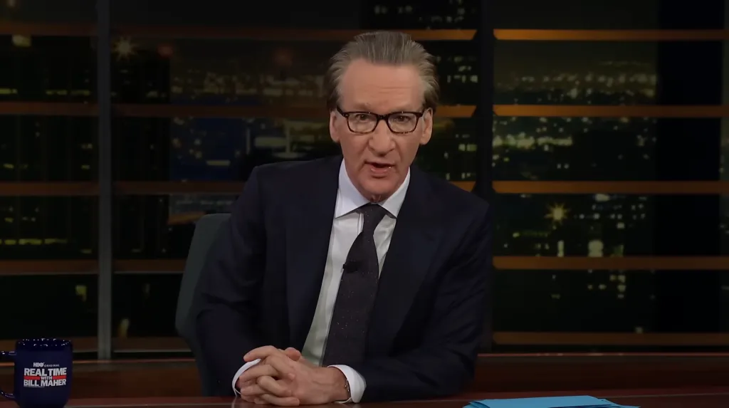 Bill Maher at a desk with a city skyline in the background.