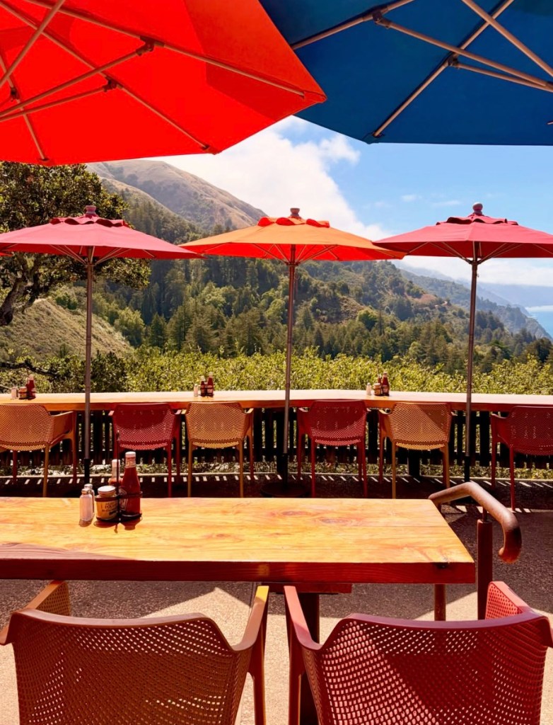 Outdoor dining area at Nepenthe in Big Sur with colorful umbrellas and a scenic view of mountains and the ocean.