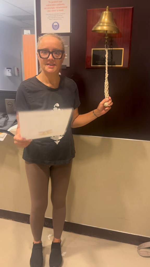 A woman in a clinic holds a paper and a bell rope, ready to ring a brass bell on a plaque.