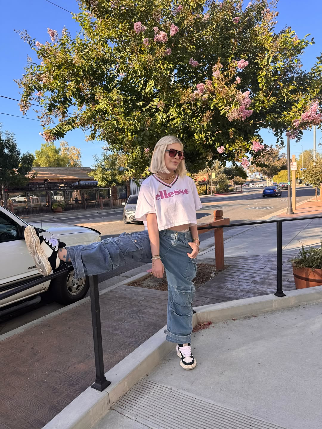 Tori Spelling posing with her leg propped on a railing, wearing a white crop top, baggy jeans, and sunglasses.