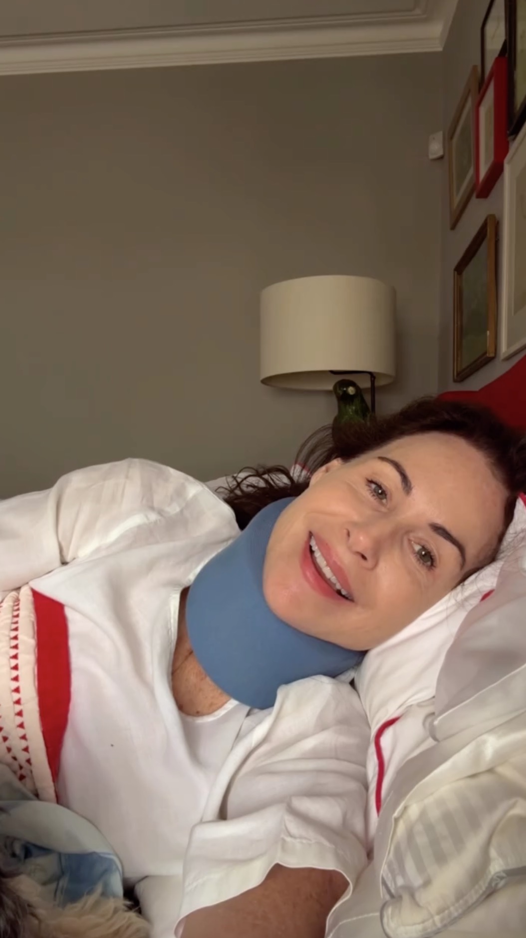 Minnie Driver in bed wearing a neck brace, with a parrot on her head.